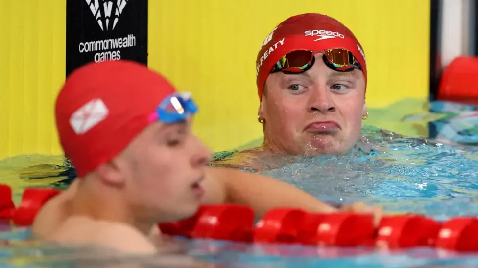 Adam Ramsay-Peaty and Filip Nowacki in the pool after their Commonwealth Games semi-final, with their heads visible above the water, both wearing red swimming caps, with goggles lifted to their foreheads.