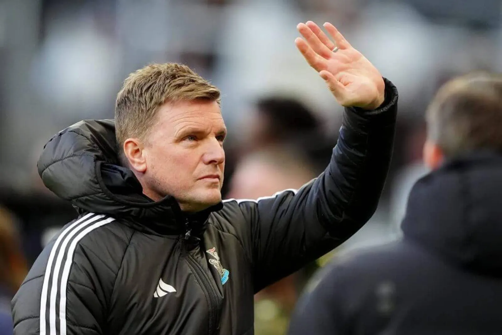 Eddie Howe, wearing a Newcastle United-branded black coat, holds up his left hand in a wave.